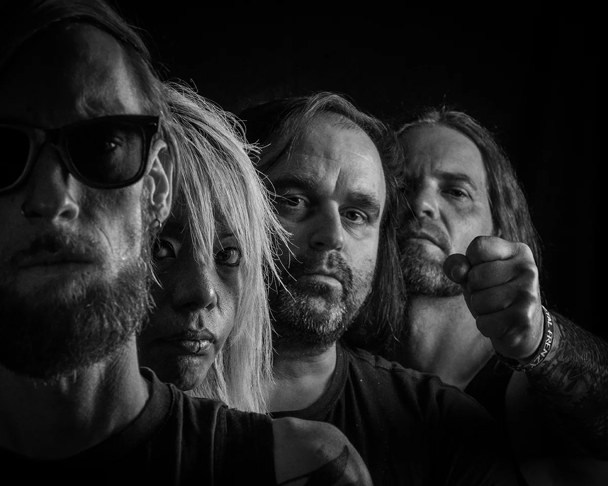 Band photo of Extinct in black and white.
Four men stand one behind the other, offset to the right. You can see them from the shoulders up. The last one stretches his fist towards the camera.