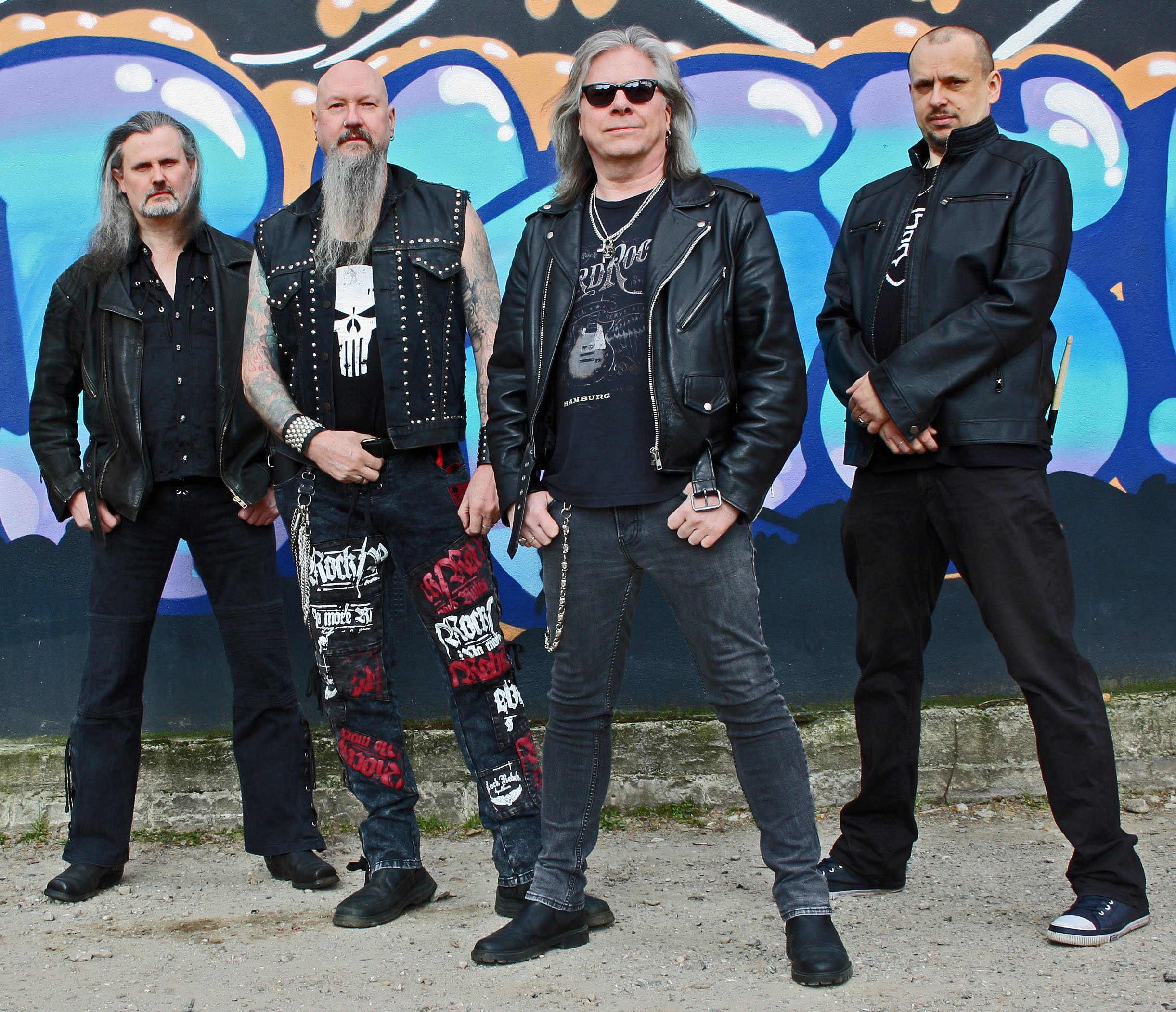 Four men, mostly dressed black with leather jackets and studs, standing in front of a graffiti wall.