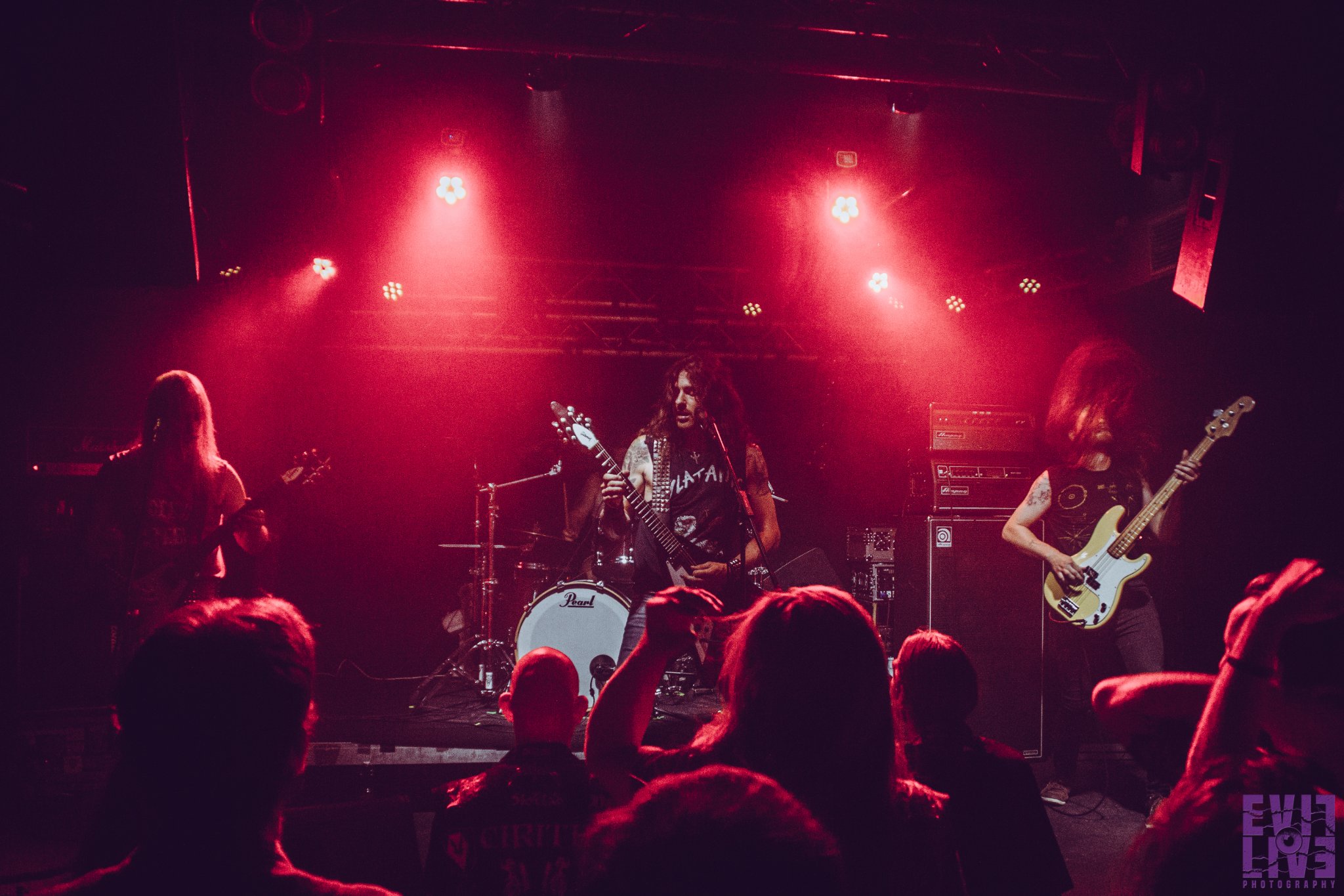 Band photo of Blitz.
Live photo from a performance. The stage is bathed in red light. Three long-haired men are in the foreground; in the background, the drum kit and drummer are only partially visible.
Guitar on the left, guitar in the middle, bass on the right.
In the foreground, the audience has their backs to the camera.