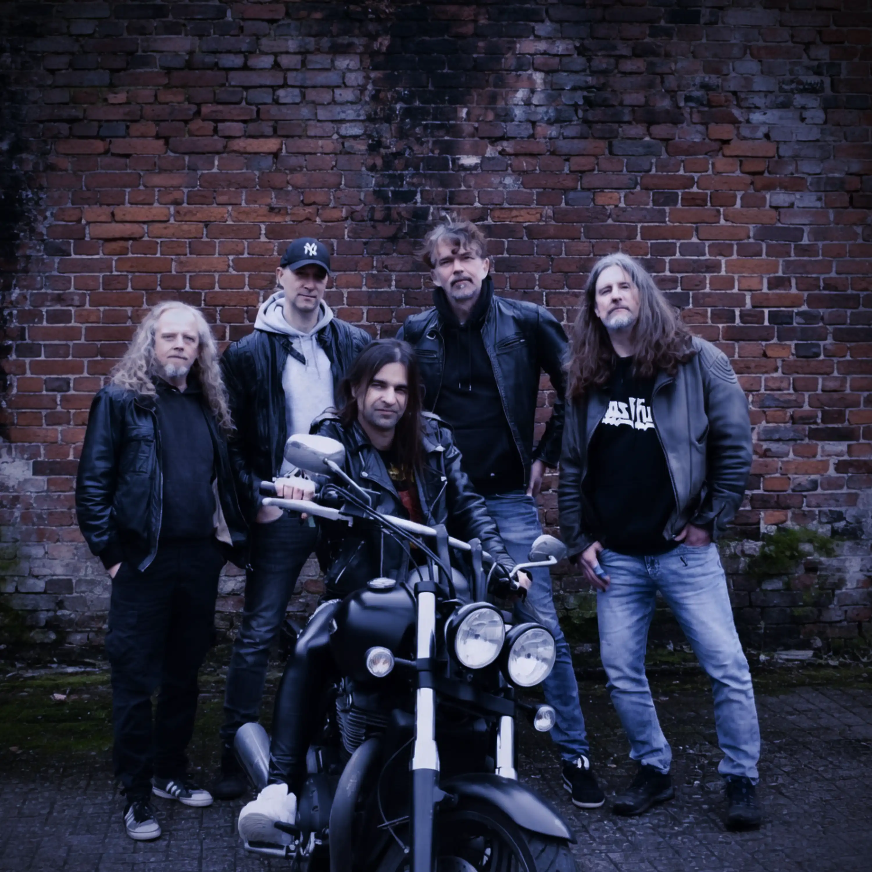 Four men in leather jackets stand in front of a partially moss-covered red brick wall; a fifth man, also in a leather jacket, sits on a black motorcycle in front of them. They look into the camera.