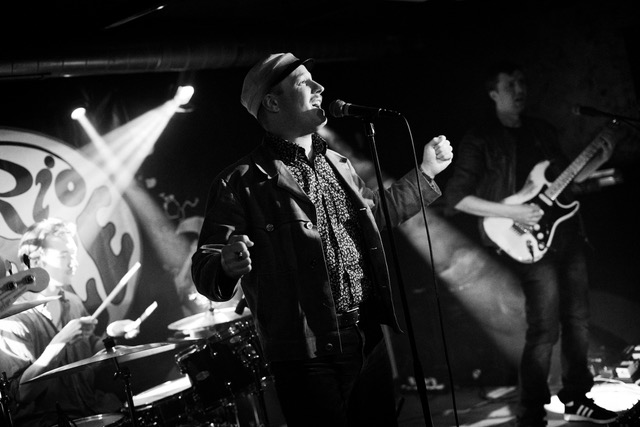 Bandfoto von Furio Grace in schwarz/weiß.
Im Vordergrund auf der Bühne steht der Sänger vor dem Mikrofon und performed.
Im Hintergrund links der Drummer hinter dem Schlagzeug, ein Spotlight auf ihm. Rechts steht der Bassist.
Der Gitarrist ist nicht im Bild.