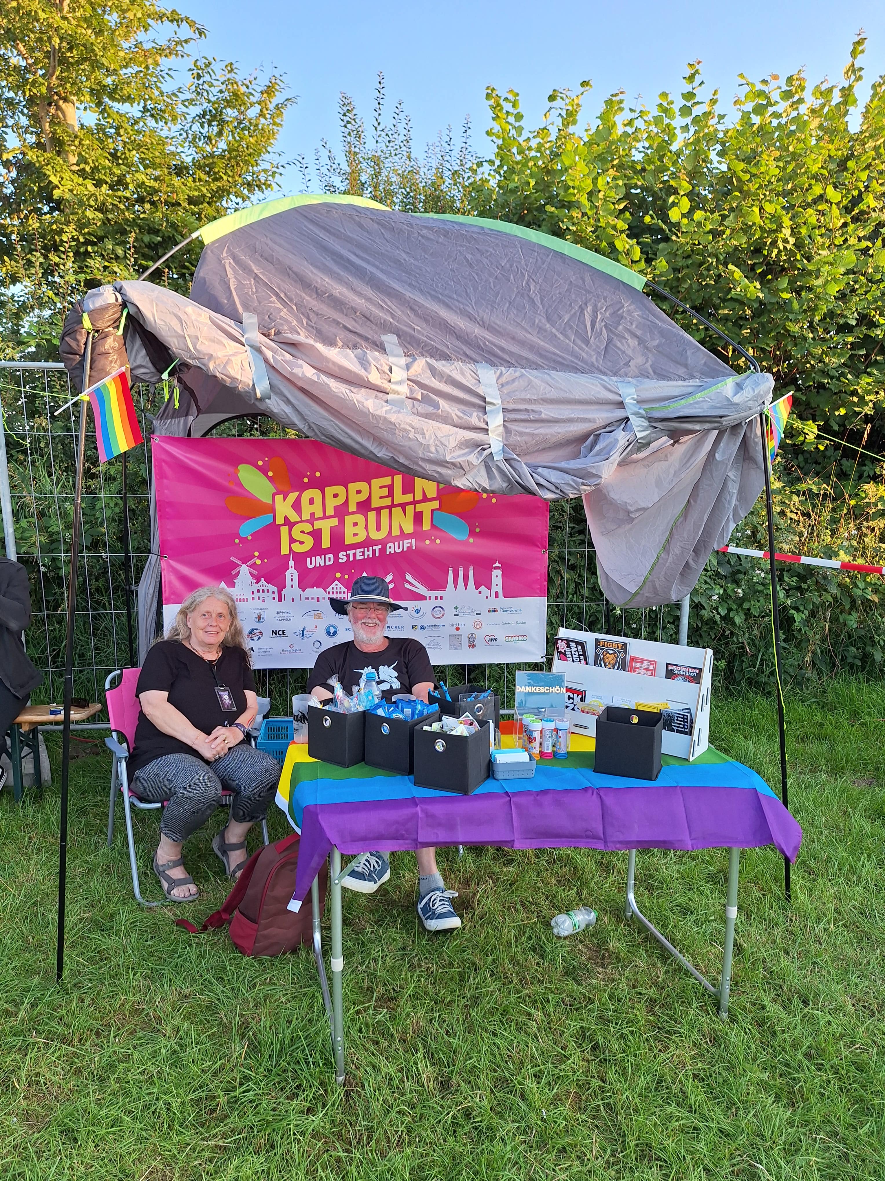 A man and a woman sit behind a table under a sunshade and smile at the camera. In the background there is a colorful banner saying "Kappeln is colorful and stands up!" stands.
The stand is decorated with rainbow flags.
On the table there are stickers, sweets, information cards and a donation box.