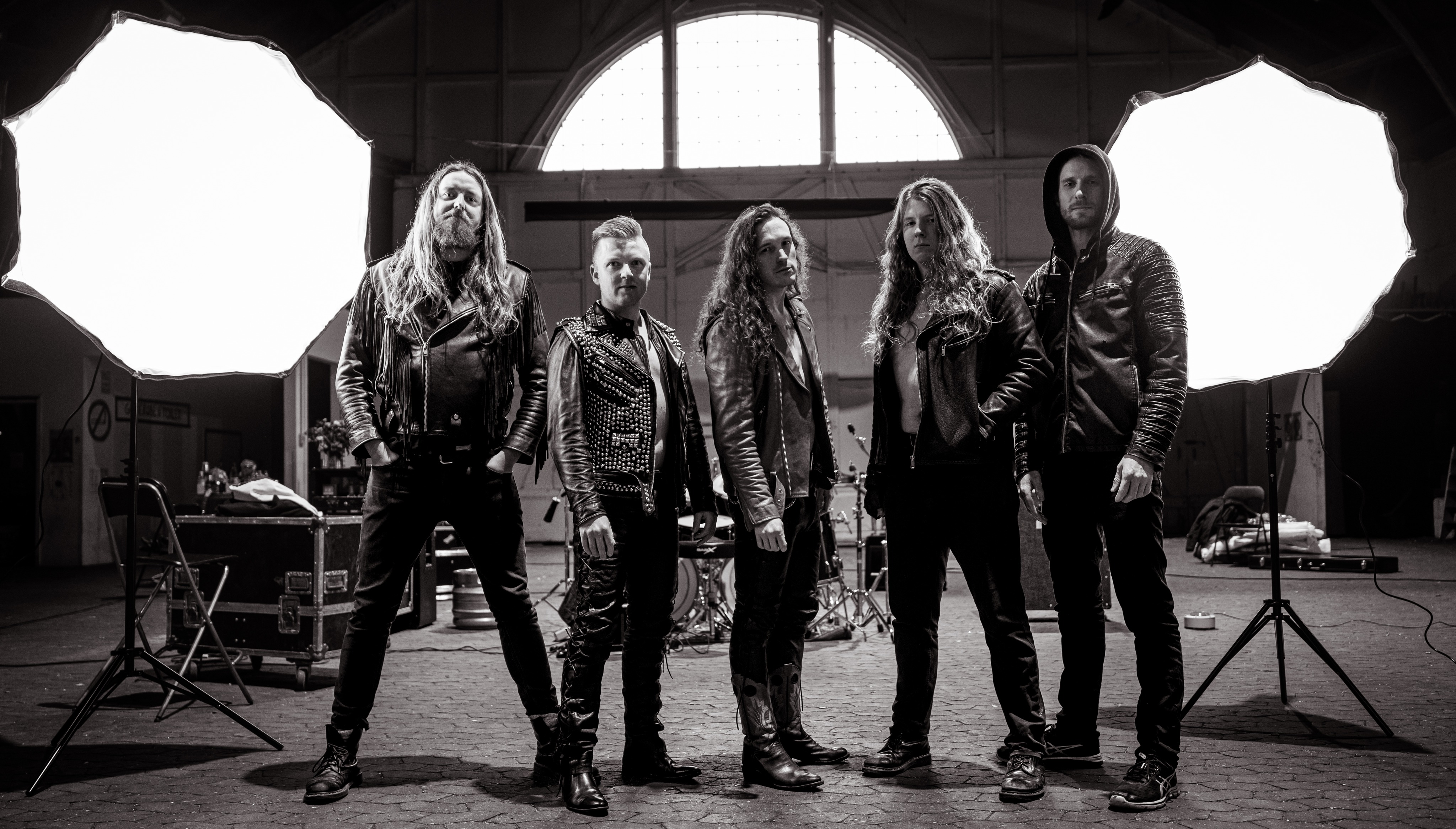 Schwarz/weißes Bandfoto von Pectora.
5 Männer stehen in einer großen  Halle, im Hintergrund steht ein Schlagzeug, Musikequipment und zwei Lightboxen.