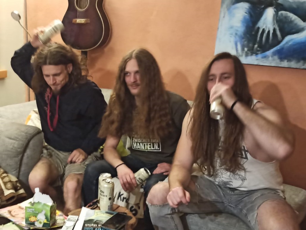 Three guys with long hair sit on a couch in the living room.
One raises his beer can over his head. One is holding his in his hand. One is drinking out of his.