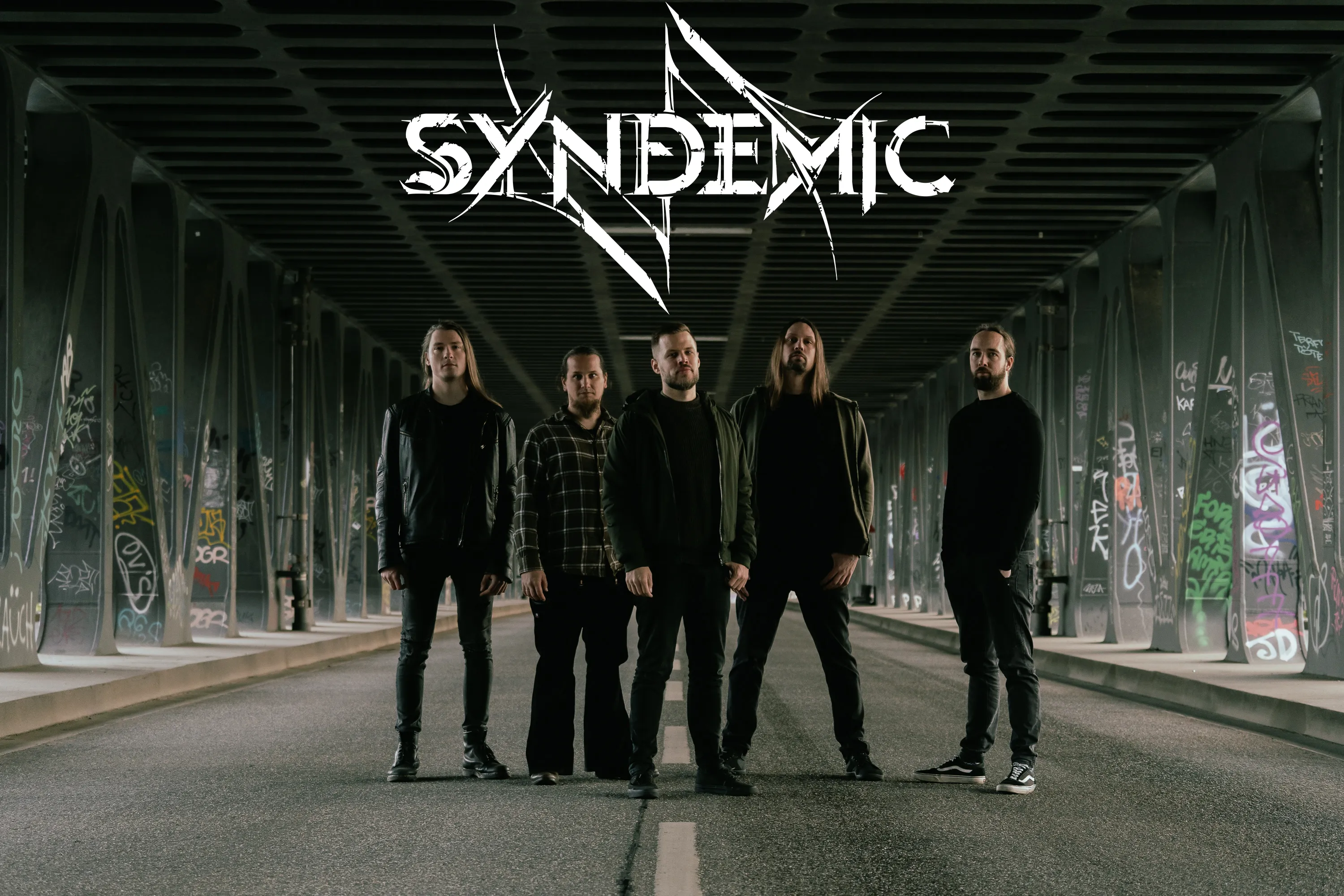 Fünf Männer stehen auf der Straße unter einer Straßenbahn. Über ihnen sieht man das Bandlogo von Syndemic.